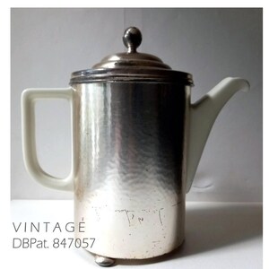 Könnte beinhalten: Eine antike Kaffeekanne aus Silber mit gehämmerter Oberfläche und einem weißen Porzellanhenkel. Die Kanne hat einen Deckel mit einem Knopf oben und einen Sockel mit drei Füßen. Der Text "DBPat. 847057" ist auf der Seite der Kanne eingraviert.