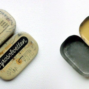 May include: Two vintage metal tins, one open and one closed. The closed tin is labeled "Tyrosolvetten" and has a yellow and white design. The open tin is labeled "T&auml;glich mehrmals 1 Pastille im Munde zergehen lassen" and has a yellow and white design.