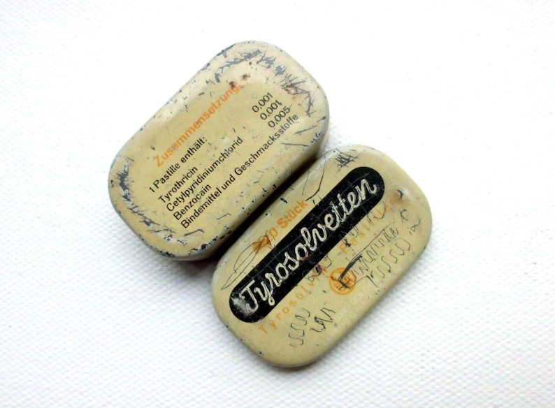 May include: Two small, rectangular, metal tins with white lids. The top tin has the text "Zusammensetzung" and a list of ingredients in German. The bottom tin has the text "Tyrosolvetten" in black lettering on a white background.