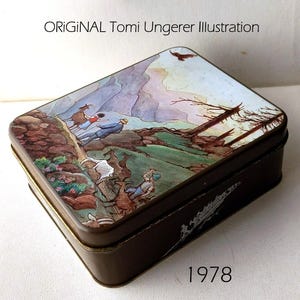 Könnte beinhalten: Eine rechteckige Metalldose mit einer farbenfrohen Illustration von Tomi Ungerer auf dem Deckel. Das Kunstwerk zeigt eine Berglandschaft mit Figuren, Ziegen und Bäumen. Die Seiten des dunkelbraunen Behälters weisen weiße grafische Details auf.