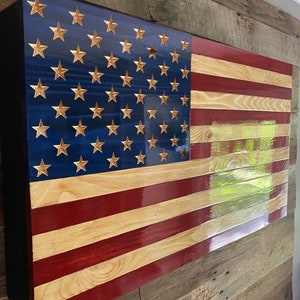 May include: A wooden American flag with red, white, and blue stripes and gold stars. The flag is mounted on a wall and has a wood grain finish.