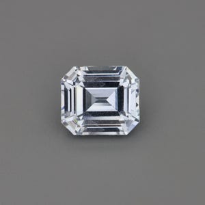 May include: A clear, emerald-cut gemstone with multiple facets. The gemstone is a bright white color and reflects light, creating a sparkling effect. The shape is rectangular with cut corners, and it is set against a gray background.