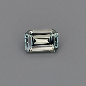 May include: A rectangular emerald-cut gemstone with a pale, icy blue-green hue. The stone features sharp, linear facets that create a clear, geometric pattern of light and dark reflections across its transparent, polished surface.