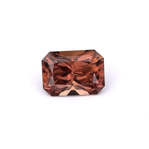 May include: A rectangular-cut gemstone with a warm, reddish-brown hue. The gem has a faceted surface that reflects light, creating a sparkling effect. The stone is set against a white background, highlighting its color and clarity.