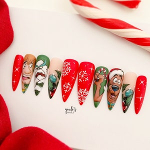 May include: Close-up of ten long, pointed, red and white Christmas-themed nail designs. Each nail features a different design, including snowflakes, holly, and characters from the cartoon Scooby-Doo. The text "giuli's NAILS" is visible at the bottom of the image.