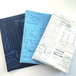 Pode incluir: Três cadernos revestidos de tecido em tons de azul. Os cadernos são impressos com designs postais e de registo vintage. O texto "POST CARD" é visível num caderno.
