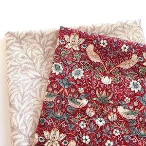 May include: Two fabric swatches. One is a light beige with a repeating leaf pattern. The other is a deep red with a floral and bird design, featuring white flowers, green leaves, and tan birds. The fabrics are folded.
