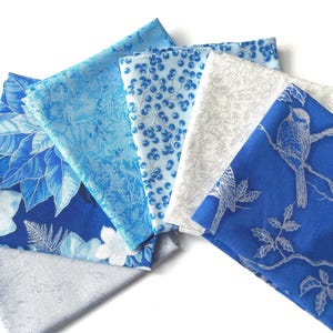 Könnte beinhalten: Eine Sammlung gefalteter Stoffquadrate in Blau- und Weißtönen. Muster umfassen Blumen, Beeren und Vogelmotive. Die Stoffe sind fächerförmig angeordnet und zeigen die Vielfalt der Texturen und Drucke.