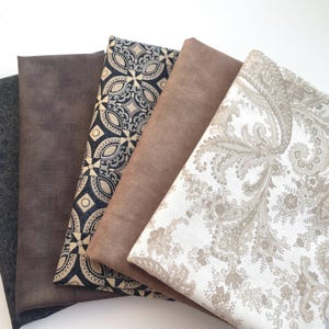 Puede incluir: Una colección de telas plegadas en varios patrones y colores. Las telas incluyen un estampado floral gris oscuro, marrones lisos y diseños estampados en negro, beige y blanco. Las telas están dispuestas en una exhibición en capas, en forma de abanico.