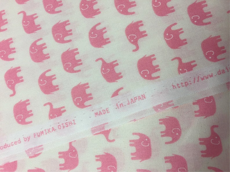 Puede incluir: Tela blanca con un patr&oacute;n repetido de elefantes rosas con caras sonrientes. El texto "MADE in JAPAN" es visible. La tela es producida por FUMIKA OISHI. La direcci&oacute;n del sitio web tambi&eacute;n es visible.