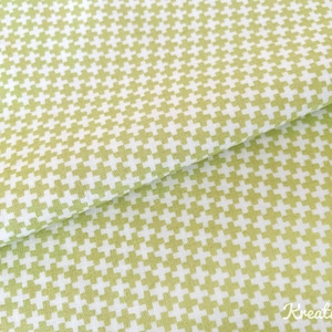 Op de afbeelding: Close-up van een gevouwen stof met een herhalend patroon van lichtgroene plustekens op een witte achtergrond. De stof heeft een zachte, getextureerde uitstraling en het patroon is gelijkmatig verdeeld. De stof is waarschijnlijk katoen.