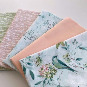 May include: A stack of five fabric pieces in various colors and patterns. The fabrics include a light green textured print, a pink and white patterned fabric, a light blue fabric with text, a solid peach fabric, and a fabric with a bird and floral design.