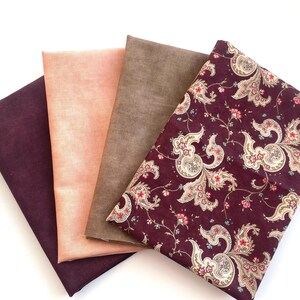 Peut inclure: Quatre échantillons de tissu dans les tons bordeaux, pêche, marron et un imprimé cachemire bordeaux. Le tissu cachemire présente un motif floral avec des accents crème, rouges et verts. Les tissus sont disposés sur une surface blanche.