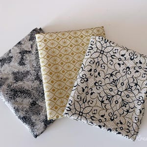 Puede incluir: Tres cuadrados de tela doblados con diferentes patrones. Uno tiene un diseño abstracto en blanco y negro, otro un patrón de estrella dorada y blanca, y el tercero presenta un diseño floral en blanco y negro.