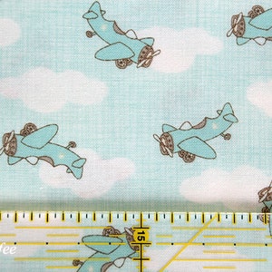 May include: Light blue fabric with a repeating pattern of cartoon airplanes and white clouds. The airplanes are teal with brown accents. A yellow ruler with black markings is at the bottom of the image.