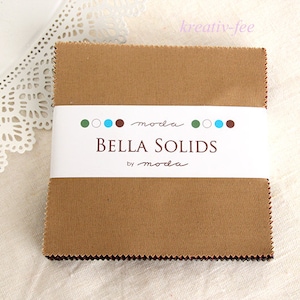 Könnte beinhalten: Ein Stapel brauner Stoffquadrate, beschriftet mit "BELLA SOLIDS by moda", ist mit einem weißen Band umwickelt. Der Stoff hat eine strukturierte Optik. Der Hintergrund ist eine weiße, spitzenartige Oberfläche.