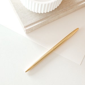 Könnte beinhalten: Eine weiße Keramikschüssel mit geripptem Design liegt auf einem beigefarbenen, mit Leinen bezogenen Notizbuch. Ein goldener Stift liegt auf einer weißen Oberfläche.