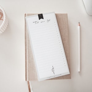 Könnte beinhalten: Ein weißer Notizblock mit einem beigefarbenen Leinenumschlag und einem weißen Bleistift. Der Notizblock hat eine schwarze Lasche mit dem Text "Alles hat seine Zeit" darauf geschrieben. Der Notizblock liegt auf einer weißen Oberfläche.