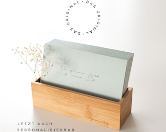 Könnte beinhalten: Ein Stapel hellblauer Karten mit dem Text "Die geheime Zeit ist immer jetzt" in einer Holzkiste. Die Kiste ist mit "grimmswald" beschriftet und der Text "JETZT AUCH PERSONALISIERBAR" befindet sich unter der Kiste.