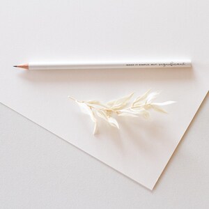 Könnte beinhalten: Ein weißer Bleistift mit dem Text "Make it simple, but significant" darauf gedruckt. Der Bleistift liegt auf einer weißen Oberfläche mit einem getrockneten Zweig weißer Blumen.