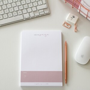 Könnte beinhalten: Ein weißer Notizblock mit einem rosa unteren Rand und dem Text "Turn the page" in kursivschrift. Der Notizblock liegt auf einer weißen Oberfläche mit einer weißen Tastatur, einer weißen Maus und einem roségoldenen Stift.
