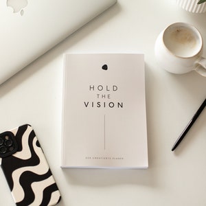 Könnte beinhalten: Ein weißer Planer mit dem Text "HOLD THE VISION" und "DER UNDATIERT PLANER" auf dem Cover. Der Planer liegt auf einer weißen Oberfläche mit einer schwarz-weißen Handyhülle, einer Tasse Kaffee, einem Stift und einer Pflanze.