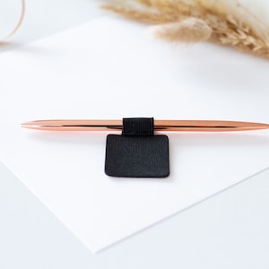 Könnte beinhalten: Ein roségoldener Stift mit einem schwarzen Stifthalter. Der Stift liegt auf einer weißen Oberfläche. Der Stifthalter ist mit einem schwarzen Gummiband am Stift befestigt. Der Hintergrund umfasst getrocknete Pflanzen.