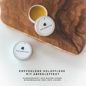 May include: Open tin of wood wax with a light brown, waxy substance, next to two closed tins. The tins have a silver rim and a white label with the text "HOLZWACHS" and "AUS NAT&Uuml;RLICHEM BIENENWACHS". The text "EMPFOHLENE HOLZPFLEGE MIT ABPERLEFFEKT" is below.