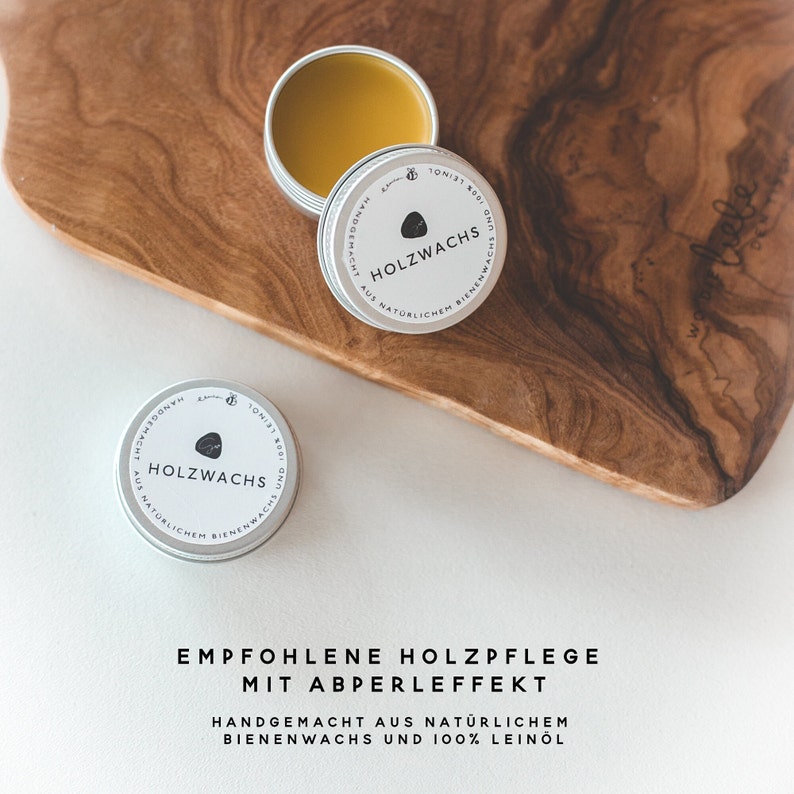 May include: Open tin of wood wax with a light brown, waxy substance, next to two closed tins. The tins have a silver rim and a white label with the text "HOLZWACHS" and "AUS NAT&Uuml;RLICHEM BIENENWACHS". The text "EMPFOHLENE HOLZPFLEGE MIT ABPERLEFFEKT" is below.