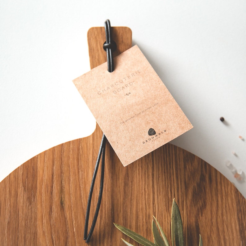 May include: A wooden charcuterie board with a handle and a black leather loop. A brown paper tag is attached with the words "CHARCUTERIE BOARD" printed on it. The board is made of light brown wood with visible grain.