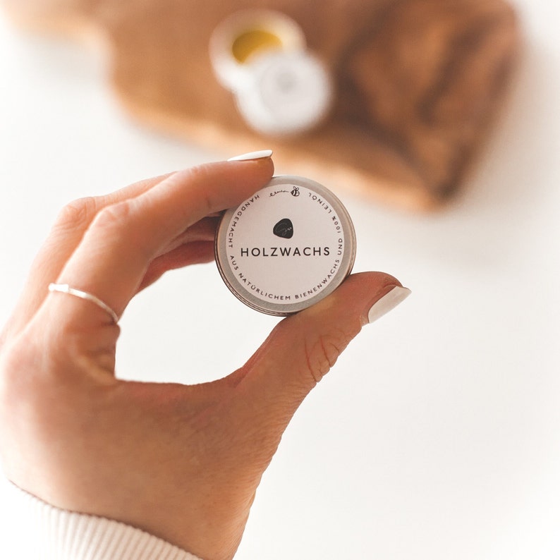 May include: A small, round tin of wood wax held in a hand. The tin is silver with black text that reads "HOLZWACHS" and "AUS NAT&Uuml;RLICHEM BIENENWACHS". The background is blurred, showing a wooden surface and another container.