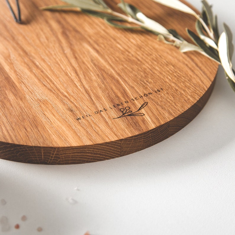 May include: A round wooden serving board with a natural wood grain finish. The board has a dark brown edge and features the text "WEIL DAS LEBEN SCHON IST" with a floral design. A sprig of greenery rests on the board.