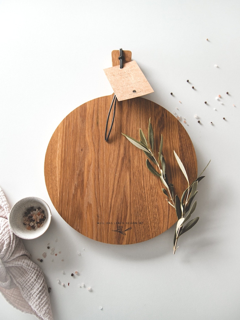 May include: A round wooden cutting board with a natural wood grain finish. The board has a handle and a tag attached. A sprig of greenery and a small bowl with salt and pepper are next to the board.