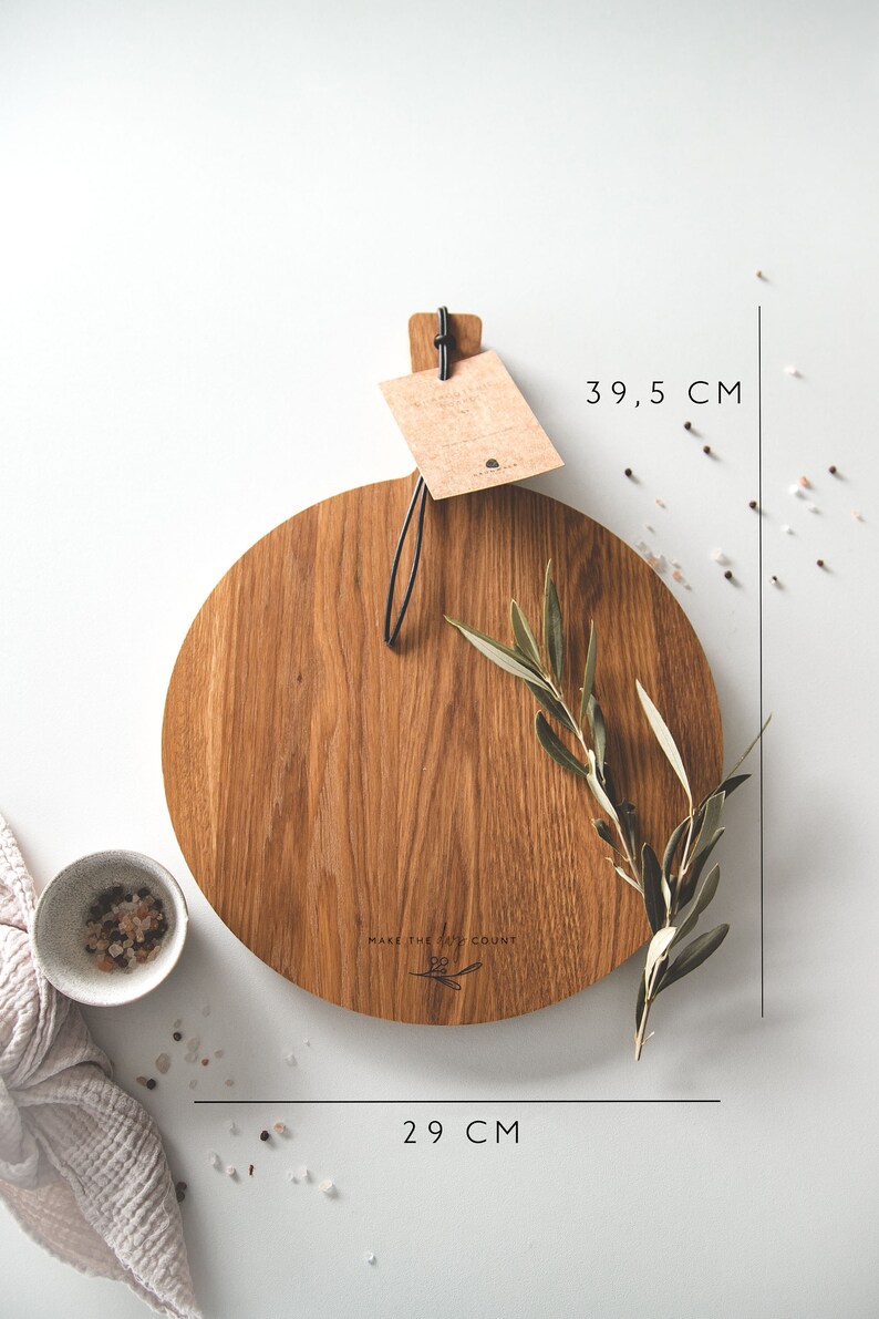 May include: A round wooden cutting board, 29 cm in diameter and 39.5 cm long, with a handle and a tag. The board has the text "MAKE THE MOMENT COUNT". An olive branch and a small bowl of salt are also present.