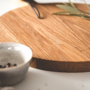 May include: A round wooden cutting board with a handle, featuring a natural wood grain. A small, speckled gray bowl with peppercorns sits in the foreground. The board has the text "WEIL DESIGN" etched on the side.
