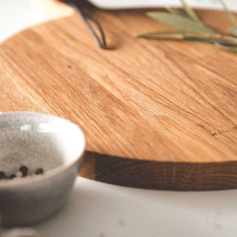 May include: A round wooden cutting board with a handle, featuring a natural wood grain. A small, speckled gray bowl with peppercorns sits in the foreground. The board has the text "WEIL DESIGN" etched on the side.