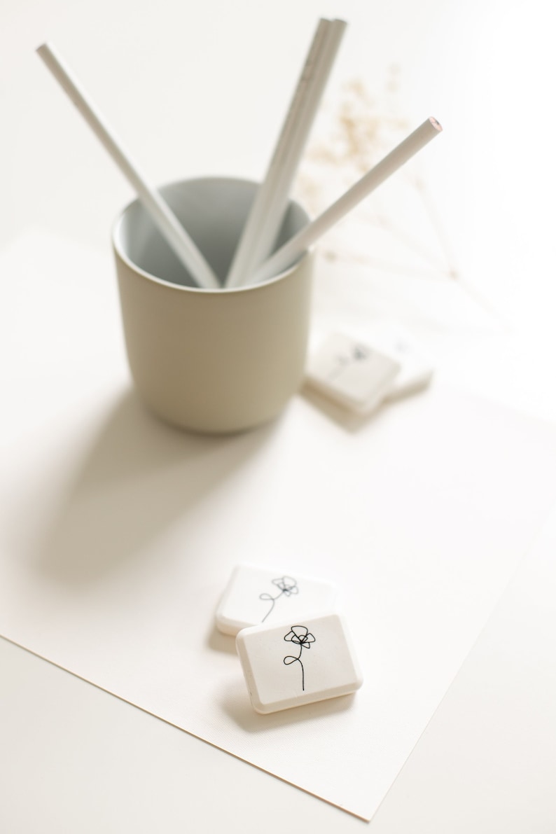 May include: Two white square stamps with a simple line drawing of a flower on each. The stamps are on a white surface next to a beige ceramic cup with white sticks inside.
