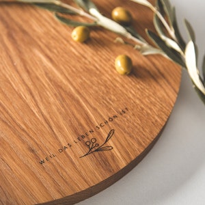 May include: A round wooden serving board with a natural wood grain finish. The board features the engraved text "WEIL DAS LEBEN SCH&Ouml;N IST" and a botanical illustration. Green olives and olive branches are arranged on the board.