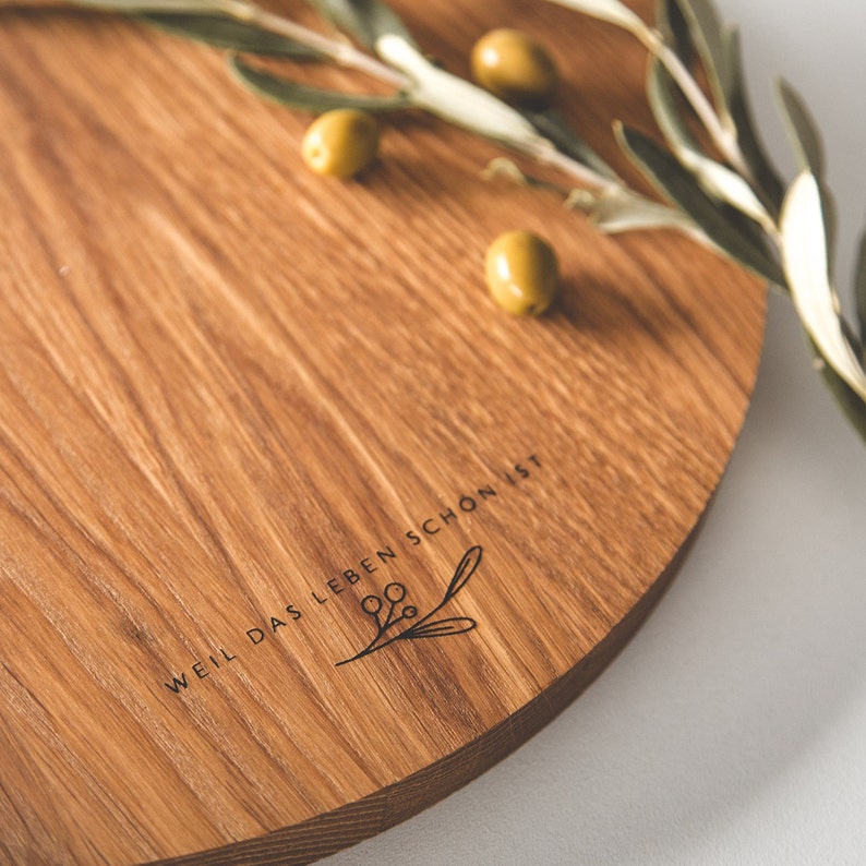 May include: A round wooden serving board with a natural wood grain finish. The board features the engraved text "WEIL DAS LEBEN SCH&Ouml;N IST" and a botanical illustration. Green olives and olive branches are arranged on the board.