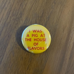 Puede incluir: Un botón amarillo con el texto "I WAS A PIG AT THE HOUSE OF FLAVORS" en rojo. El botón es redondo y tiene un acabado brillante. El fondo es una superficie de madera.