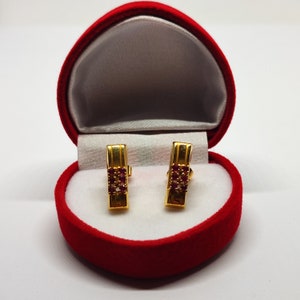 Könnte beinhalten: Ein Paar goldene Ohrringe mit roten Edelsteinen, die in einer rechteckigen Form gefasst sind, in einer roten Samt-Schmuckschatulle präsentiert.