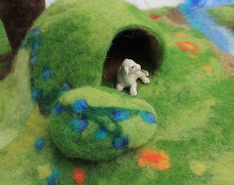 May include: A white lamb figurine sits inside a green felt cave with blue and green flowers. The cave is on a green felt surface with a blue felt stream.