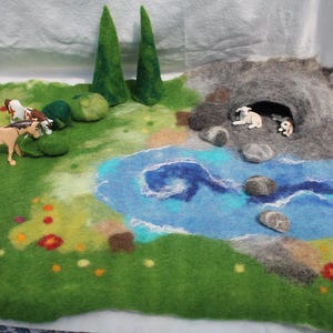 Könnte beinhalten: Eine gefilzte Landschaftsszene mit einer grünen Wiese, blauem Wasser und einer grauen Höhle. Miniaturpferde und -schafe sind auf der Landschaft platziert. Zwei grüne Filzbäume und bunte Blumen ergänzen die Szene.