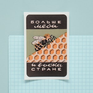 Könnte beinhalten: Ein Aufkleber im Vintage-Stil mit einer Schwarzweißillustration einer Biene auf einer Wabe. Der Text auf dem Aufkleber lautet "Больше меда и воска в стране".