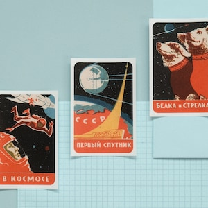 Puede incluir: Tres pegatinas de temática espacial de estilo vintage. Una presenta a un astronauta con un traje espacial rojo con el texto "B KOCMOCE". Otra muestra un cohete lanzándose con el texto "ПEPBЫЙ CПYTHИK". La última representa a dos perros en el espacio con el texto "БEЛKA И CTPEЛKA".