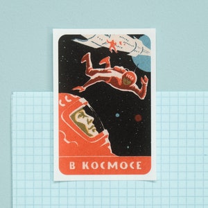 May include: Vintage-style sticker featuring a retro space scene. An astronaut in a red spacesuit floats in space near a spaceship. The sticker has the Russian text "B KOCMOCE" at the bottom, meaning "In Space".