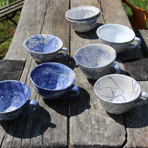 Puede incluir: Una colecci&oacute;n de siete tazas de t&eacute; de cer&aacute;mica azul y blanca con un acabado de esmalte craquelado. Las tazas est&aacute;n dispuestas sobre una mesa de madera.