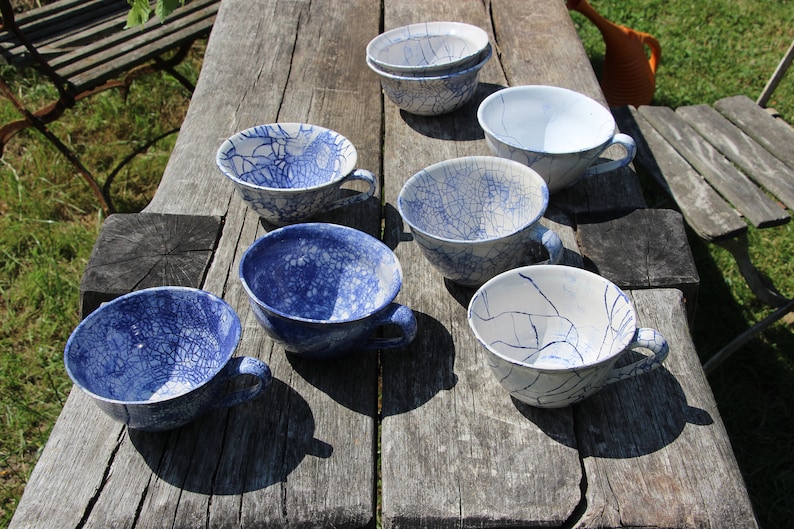 Puede incluir: Una colecci&oacute;n de siete tazas de t&eacute; de cer&aacute;mica azul y blanca con un acabado de esmalte craquelado. Las tazas est&aacute;n dispuestas sobre una mesa de madera.