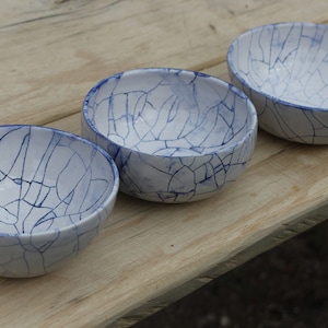 May include: Three white ceramic bowls with a blue crackle glaze pattern. The bowls are arranged in a row on a wooden surface.
