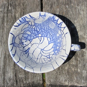 Puede incluir: Una taza de t&eacute; de cer&aacute;mica blanca con un esmalte craquelado azul. La taza tiene un asa y est&aacute; sentada sobre una superficie de madera.
